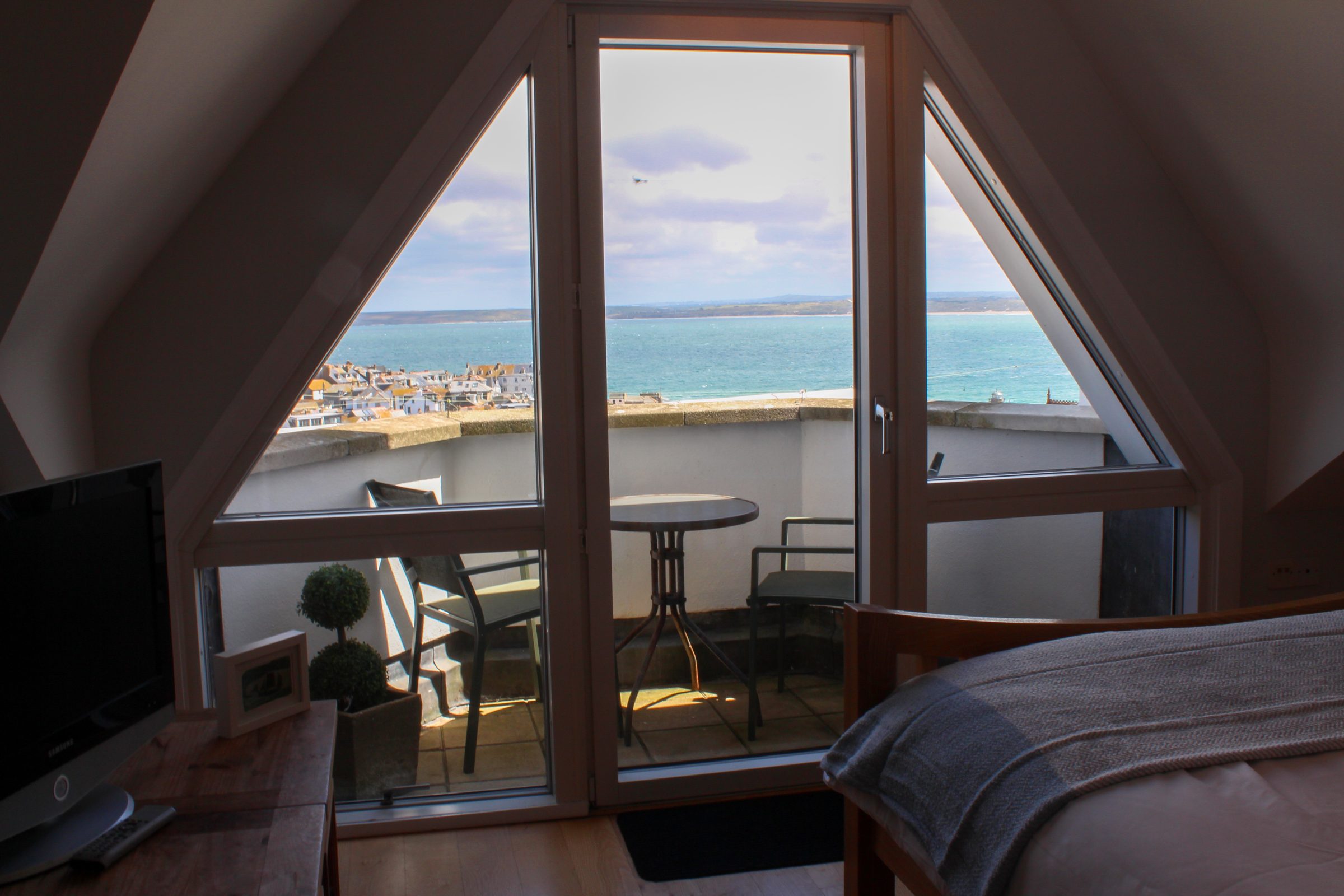 The apex bedroom window at Tate Five, framing the balcony, St Ives rooftops and the bay beyond.