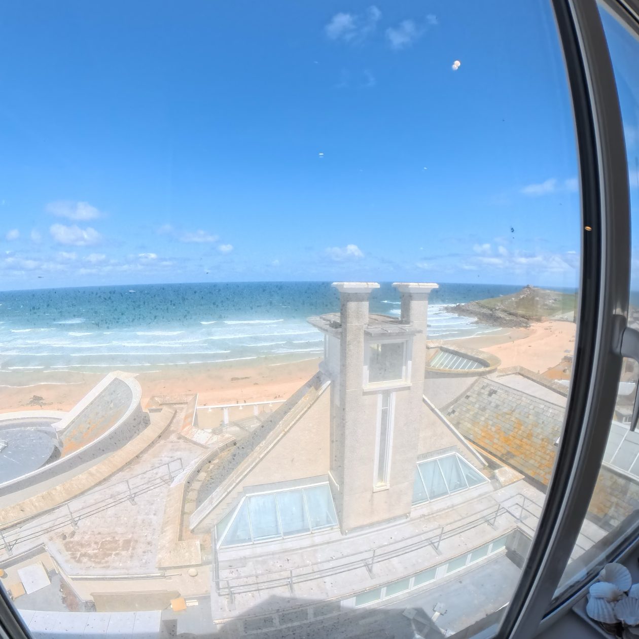 Looking down through the landing's porthole window onto the roof lanterns of Tate St Ives, with Porthmeor Beach and the surf beyond.